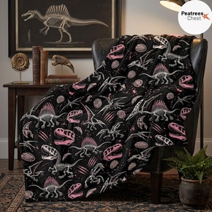 May include: A black throw blanket featuring a repeating pattern of white dinosaur skeletons and pink fossilized skulls. The design includes various prehistoric creatures, including winged pterosaurs and spined dinosaurs, scattered across the fabric.
