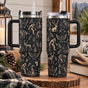 May include: Two black stainless steel tumblers with handles and clear lids featuring a repeating gold-toned pattern of cryptids, skeletons, forest elements, and crescent moons. Each tumbler includes a clear straw and a tapered base design.