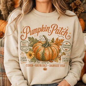 Könnte beinhalten: Ein beigefarbenes Sweatshirt mit Rundhalsausschnitt und einer großen Grafik eines orangefarbenen Kürbisses, umgeben von Blättern und Blumen. Der Text lautet Pumpkin Patch, Cozy Fall Harvest, Heirloom Pumpkins, Farm Stand, Locally Grown, Cider, Hayrides, Harvest Fest.