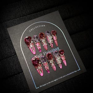 May include: A set of ten long, pink glittered artificial nails. Each nail features a large, jewel-toned gemstone and intricate silver embellishments. The nails are displayed on a black card with the text "Nail Painting" and "Collection."