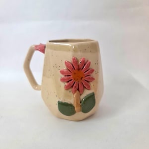 Può includere: Una tazza in ceramica beige con finitura maculata, caratterizzata da un fiore rosa tridimensionale in rilievo con centro arancione e due foglie verdi sul davanti. La tazza presenta un manico angolare con un piccolo accento decorativo rosa.