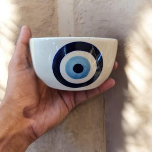 May include: A small, off-white ceramic bowl featuring a hand-painted evil eye motif on the front. The design consists of a dark blue outer ring, a light blue middle circle, and a solid black center pupil, creating a distinct circular pattern.