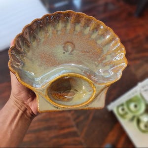 Può includere: Un piatto in ceramica a forma di conchiglia con una smaltatura lucida nei toni del marrone, dell'abbronzatura e del verde tenue. Il piatto presenta un bordo smerlato e un piccolo scomparto ovale incassato alla base, con una texture maculata.