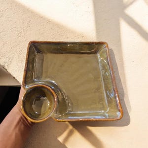 Könnte beinhalten: Eine quadratische Servierschale aus Keramik mit einer glänzenden olivgrünen Glasur und einem dunkelbraunen Rand. Die Schale verfügt über eine ausgeprägte kreisförmige Vertiefung in einer Ecke, die als kleine integrierte Schale für Dips dient.