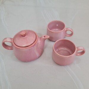 May include: A ceramic tea set consisting of a small teapot with a matching lid and two rounded cups, all finished in a uniform glossy pink glaze. The teapot features a curved handle and spout, while each cup has a single side handle.