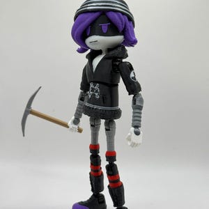 May include: A posable action figure with purple hair, a black beanie with white stripes, and a black hooded jacket featuring a white graphic. The figure has grey robotic limbs, red-striped leg accents, black shoes with purple toes, and holds a pickaxe.
