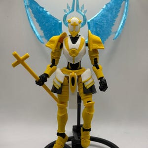 Könnte beinhalten: Eine gelb-weiße Actionfigur eines Ritters mit blauen Flügeln und einem kreuzförmigen Stab. Die Figur hat einen Helm, Schulterpolster und ein rockartiges Teil. Sie steht auf einem schwarzen, kreisförmigen Sockel.
