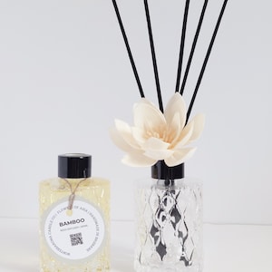 May include: Two glass reed diffusers. One contains a yellow liquid and a label that reads "BAMBOO REED DIFFUSER". The other is clear with a decorative flower and black reeds. The bottles have a diamond pattern.