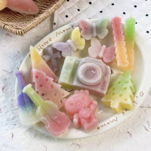 Puede incluir: Una colección de figuras de cera en tonos pastel sobre un plato blanco con el texto Home Moment. Las formas incluyen una cámara, mariposas, árboles de Navidad, guitarras y una figura de gato, con degradados suaves de rosa, amarillo, verde y morado.