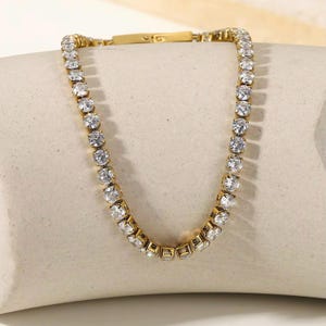 May include: A gold-toned tennis necklace featuring a series of clear, round-cut cubic zirconia stones. The necklace has a classic design with a secure clasp. The stones are set closely together, creating a continuous sparkle. This piece is suitable for various occasions.