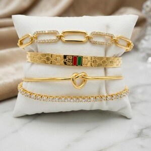 May include: A set of four gold-toned bracelets displayed on a white cushion. The top bracelet is a chain link with crystals, the second is a patterned cuff with red and green enamel, the third is a thin band with a heart knot, and the bottom is a crystal tennis bracelet.
