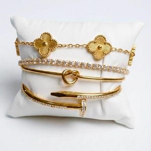 May include: A set of four gold-toned bracelets displayed on a white cushion. The collection includes a chain bracelet with two clover-shaped charms, a crystal-encrusted tennis bracelet, a smooth bangle with a knot detail, and a nail-shaped bangle.