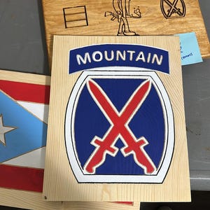 May include: A square wooden plaque featuring the 10th Mountain Division insignia, displaying a blue shield with two crossed red swords, topped by a blue arch with the white text MOUNTAIN. The design is painted onto a light-colored wood board.