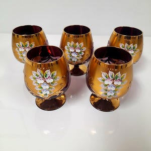 Puede incluir: Un juego de cinco copas de cristal de color rojo intenso con un acabado dorado metálico en el exterior. Cada copa está decorada con un motivo floral pintado a mano en blanco, azul y verde, con detalles dorados y una forma redondeada.