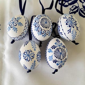 Op de afbeelding: Vijf witte eivormige ornamenten versierd met ingewikkelde blauwe bloemmotieven en geometrische patronen. Elk ornament is voorzien van een donkerblauw lintlusje aan de bovenkant om op te hangen, met gedetailleerde handgeschilderde ontwerpen.