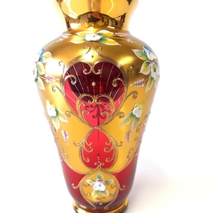 May include: A glass vase featuring a deep red base color with an overlay of metallic gold paint. The surface is decorated with hand-painted white and green floral motifs and intricate gold filigree scrollwork patterns across the exterior.