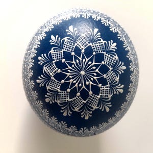 May include: A dark blue egg decorated with intricate white hand-painted patterns. The design features a central star-like mandala motif surrounded by geometric grid shapes and delicate floral-inspired borders along the outer edge of the surface.