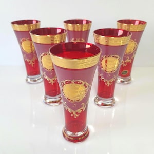 Può includere: Un set di sei bicchieri alti in vetro rosso rubino con un'ampia fascia dorata sul bordo e dettagli in filigrana d'oro. Ogni bicchiere presenta un emblema centrale dorato con la scritta Bohemia sulla superficie in vetro rosso intenso.