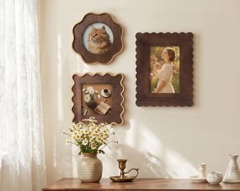 May include: Three dark brown wooden picture frames with scalloped edges mounted on a wall. One frame is circular, one is square, and one is rectangular. The frames contain photographs of a cat, a still life with flowers, and a person in a field.