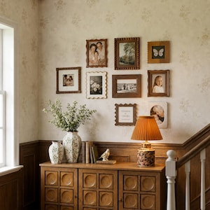 Könnte beinhalten: Eine Galeriewand mit neun verschiedenen rechteckigen und quadratischen Bilderrahmen in Holz- und Cremetönen, die verschiedene Fotos und Kunstwerke zeigen. Darunter steht ein Holzschrank mit zwei Blumenvasen, Büchern, einer Vogelfigur und einer Plisseelampe.