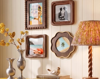 May include: Four wooden wall frames with varied shapes including a beaded rectangular frame, a square frame, a wavy square frame, and a flower-shaped frame, each displaying a different photograph, arranged on a wall above a wooden surface.
