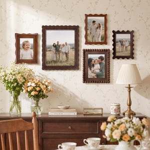 Könnte beinhalten: Eine Galerie-Wand mit fünf dunkelbraunen Bilderrahmen mit gewellten oder perlenartigen Rändern, die Familienfotos zeigen. Die Rahmen hängen an einer Wand mit Blumenmuster über einem hölzernen Sideboard mit verschiedenen Dekorationsgegenständen.