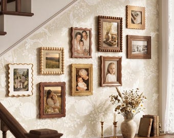 May include: A gallery wall arrangement featuring nine rectangular picture frames in various wood tones and textures, including beaded and scalloped edges. The frames display assorted photographs and are mounted on a wall with light floral wallpaper.