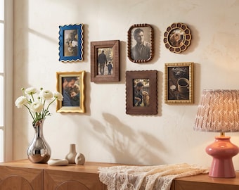 May include: A gallery wall arrangement featuring six assorted picture frames in blue, gold, and brown tones, displaying various photographs. Below, a silver vase with white flowers and a pink lamp sit on a wooden surface with a lace runner.