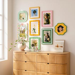 May include: A gallery wall arrangement featuring eight assorted picture frames in various shapes, sizes, and colours, including scalloped, striped, and solid designs. The frames display graduation photos and floral imagery, mounted above a light wood dresser.