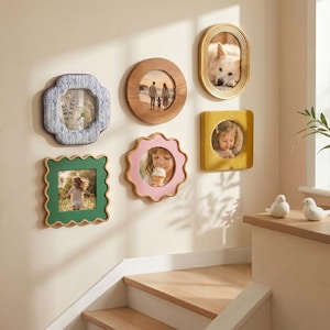 May include: A collection of decorative picture frames in various shapes and colors, mounted on a wall. The frames hold photographs and artwork. The frames include a green square, a pink scalloped frame, a wooden round frame, and a gold oval frame.