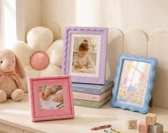 May include: Three picture frames in pink, lavender, and light blue with scalloped or beaded edges. The frames display a photograph of a child drawing, a drawing of a house and flowers, and a close-up of hands using a crayon. Includes a plush toy.