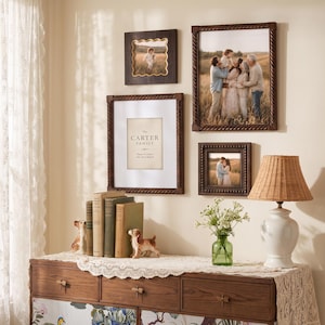 May include: A gallery wall arrangement featuring four dark wood frames with photographs and text, including a print reading The Carter Family. Below, a wooden dresser holds two dog figurines, a green glass vase with flowers, and a table lamp.