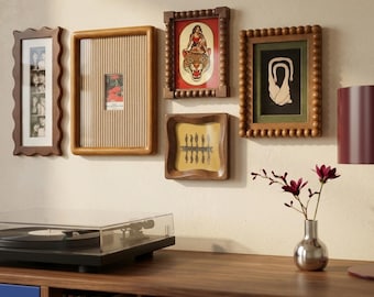 May include: A gallery wall arrangement featuring five wooden picture frames of varying shapes and textures, including scalloped, ribbed, and beaded designs, displaying diverse artwork and photographs, positioned above a record player and a vase.