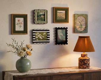 May include: A collection of six wall-mounted picture frames in various shapes and colors, including green, black, and white striped designs. A light green ceramic vase with cream and peach flowers sits next to a lamp with a pleated shade.