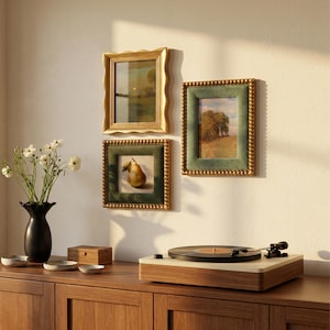 May include: Three wall-mounted picture frames with artwork, including one gold wavy frame and two green velvet frames with beaded gold edges. A black vase with white flowers, a wooden box, and a record player sit on a wooden surface.