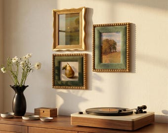 May include: Three wall-mounted picture frames with gold-toned borders, two featuring green velvet accents, displaying landscape and pear artwork. A black vase with white flowers and a record player sit on a wooden surface below.