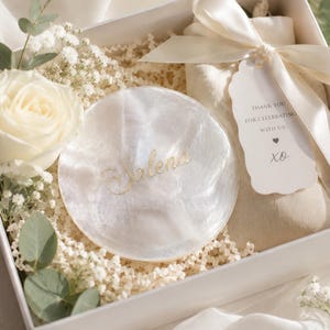 May include: A white gift box containing a round, pearlescent white trinket dish with the gold script name Valena, a beige fabric pouch tied with a cream ribbon, and a white tag reading THANK YOU FOR CELEBRATING WITH US! XO, accented by white roses.