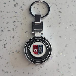 May include: A silver-colored keychain with a round emblem featuring a red and blue crest design. The emblem is surrounded by a black border with the word "ALPINA" in white lettering. The keychain has a split ring for keys.