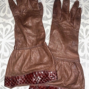 Peut inclure: Une paire de gants en cuir marron avec une finition texturée et une manchette évasée. Les poignets sont ornés d'une bordure à motif peau de serpent brun rougeâtre foncé sur le bord inférieur, offrant un contraste net avec le cuir lisse.