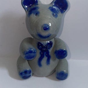 May include: A small, seated ceramic bear figurine with a glossy light grey finish. The bear features vibrant cobalt blue accents on its ears, eyes, nose, mouth, paws, and a decorative bow tie painted on its chest, creating a distinct contrast.
