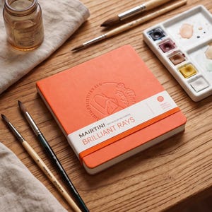 Puede incluir: Un cuaderno de bocetos de acuarela naranja con el texto "MAIRTINI BRILLIANT RAYS" y una banda blanca. La imagen también muestra pinceles, una paleta de acuarela con varios colores y un pequeño frasco de vidrio sobre una superficie de madera.