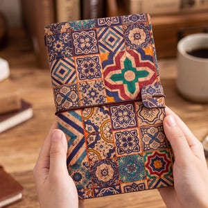 May include: A cork journal with a colorful, geometric tile pattern in shades of blue, red, green, and gold. The journal is held open, revealing the patterned cover and a brown leather strap closure.