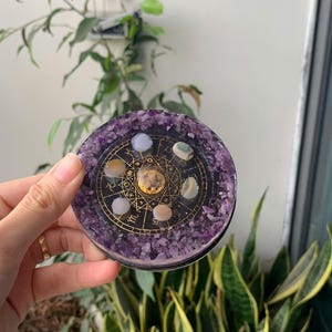 May include: A round, purple resin coaster with a gold astrological design and embedded gemstones. The coaster is surrounded by amethyst crystals and is held in front of a background of green plants.