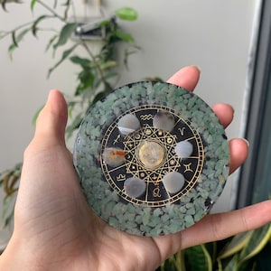 May include: Round, green resin coaster with embedded small green stones. The center features a gold-colored zodiac design with various symbols and gemstones. A unique, decorative item for home decor.