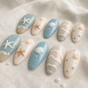 Peut inclure: Un ensemble de dix faux ongles sur le thème côtier avec une palette de couleurs bleu poussiéreux, crème et sable. Les motifs incluent des étoiles de mer et coquillages en relief 3D, des vagues blanches et des petits points bleus et blancs.