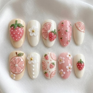 May include: A set of ten press-on nails featuring a variety of designs including raised 3D strawberries, pink citrus slices, and white flowers with gold centers. The base colors are cream and soft pink, with some nails displaying small strawberry patterns.