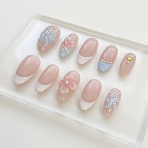 Könnte beinhalten: Ein Set aus zehn künstlichen Fingernägeln mit nudefarbener Basis und verschiedenen Designs, darunter weiße French-Spitzen, dreidimensionale rosa Blüten mit Perlen in der Mitte, hellblaue sternartige Muster und ein goldener Stern mit kleiner Perle.