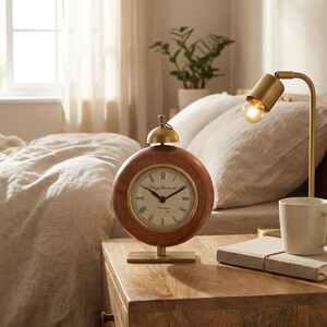 May include: A round, wooden alarm clock with a brass bell and base. The clock face has Roman numerals and the text "Village Clockworks". The clock is on a wooden nightstand next to a lamp and a mug.
