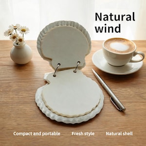 Può includere: Un blocco note a forma di conchiglia con pagine bianche rilegate da due anelli metallici. Il blocco note presenta un design con bordi smerlati testurizzati. Il testo 'Natural wind', 'Compact and portable', 'Fresh style' e 'Natural shell' è visibile.