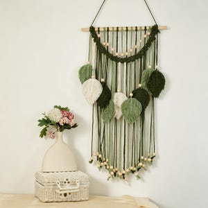 May include: A macrame wall hanging featuring a wooden dowel, various shades of green and white cotton cord, and wooden beads. The design includes multiple leaf-shaped fringe elements suspended from the dowel, creating a layered, textured appearance.